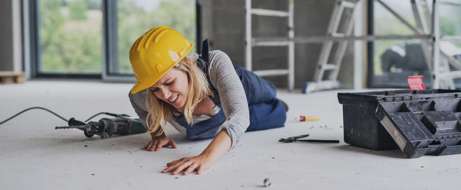 A worker in a hard hat lying injured beside a dropped drill after a construction accident